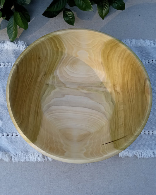 top view of a light wood  bowl with stunning grain pattern and contrasting cream and golden brown hues set on a blue woven cotton table runner with greenery at the edges