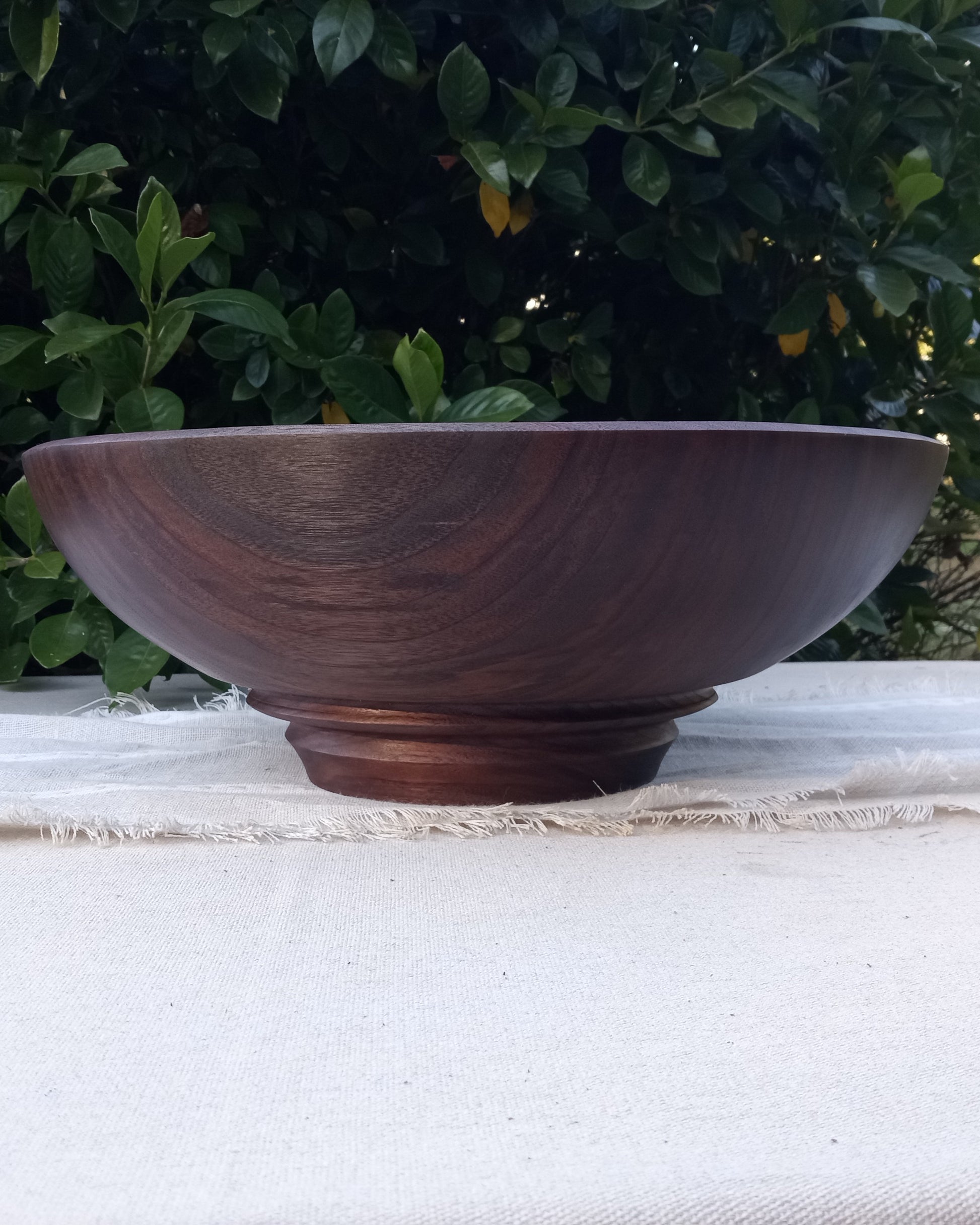 an elegant dark walnut wood bowl with a tapered round shape and an exquisitely carved pedestal base set on a white cotton table cloth