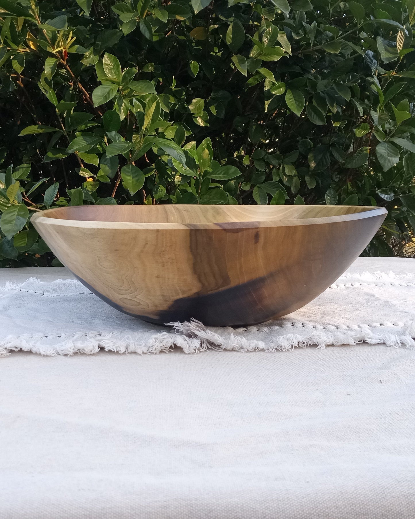 a highly figured natural edge wood bowl with stunning color contrasts of black, amber, olive and cream tones sitting on a pale blue cotton table runner with greenery in the background