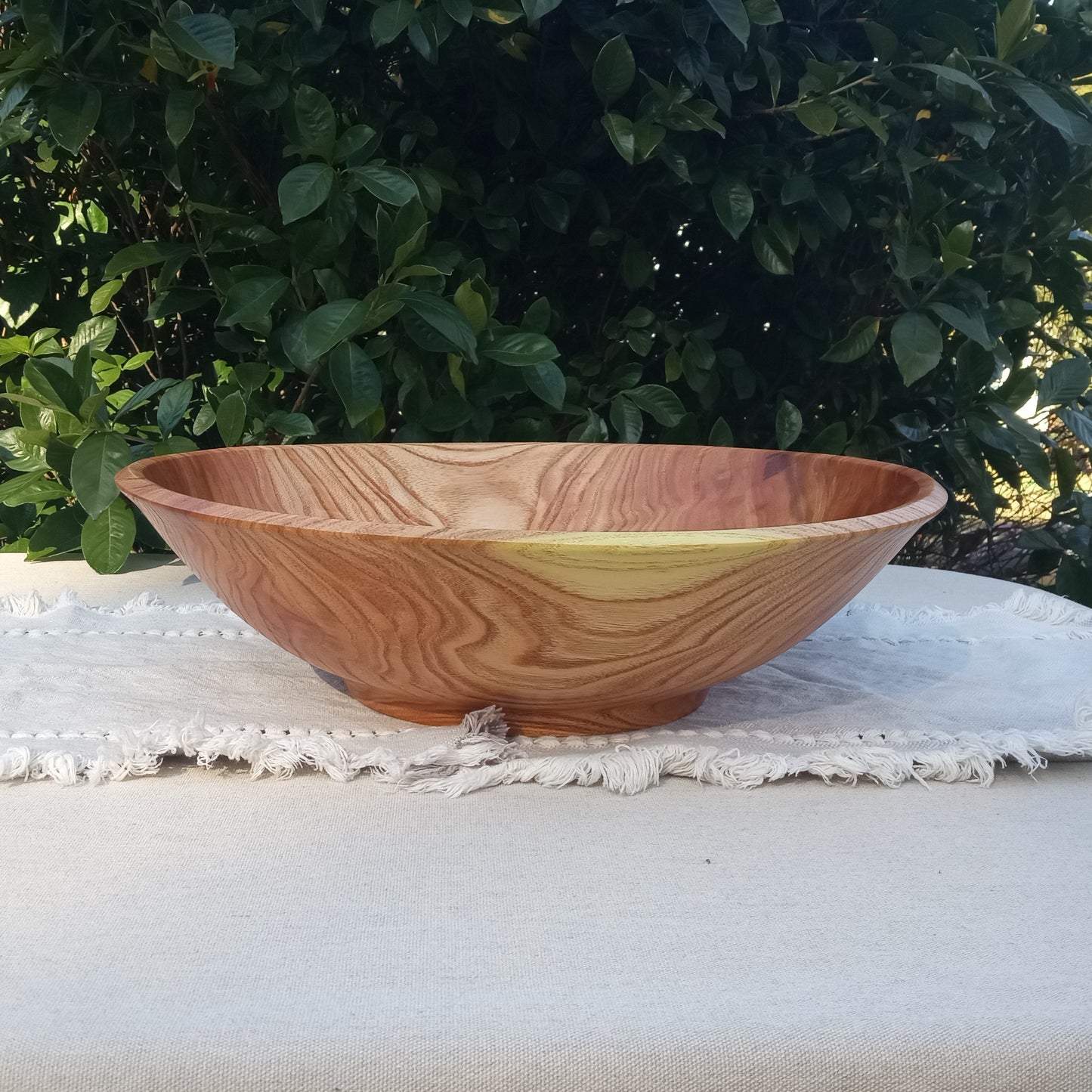 a wide round wooden bowl with striking pink and yellow hues and a wild grain pattern sitting on a white woven cotton table runner with greenery in the background