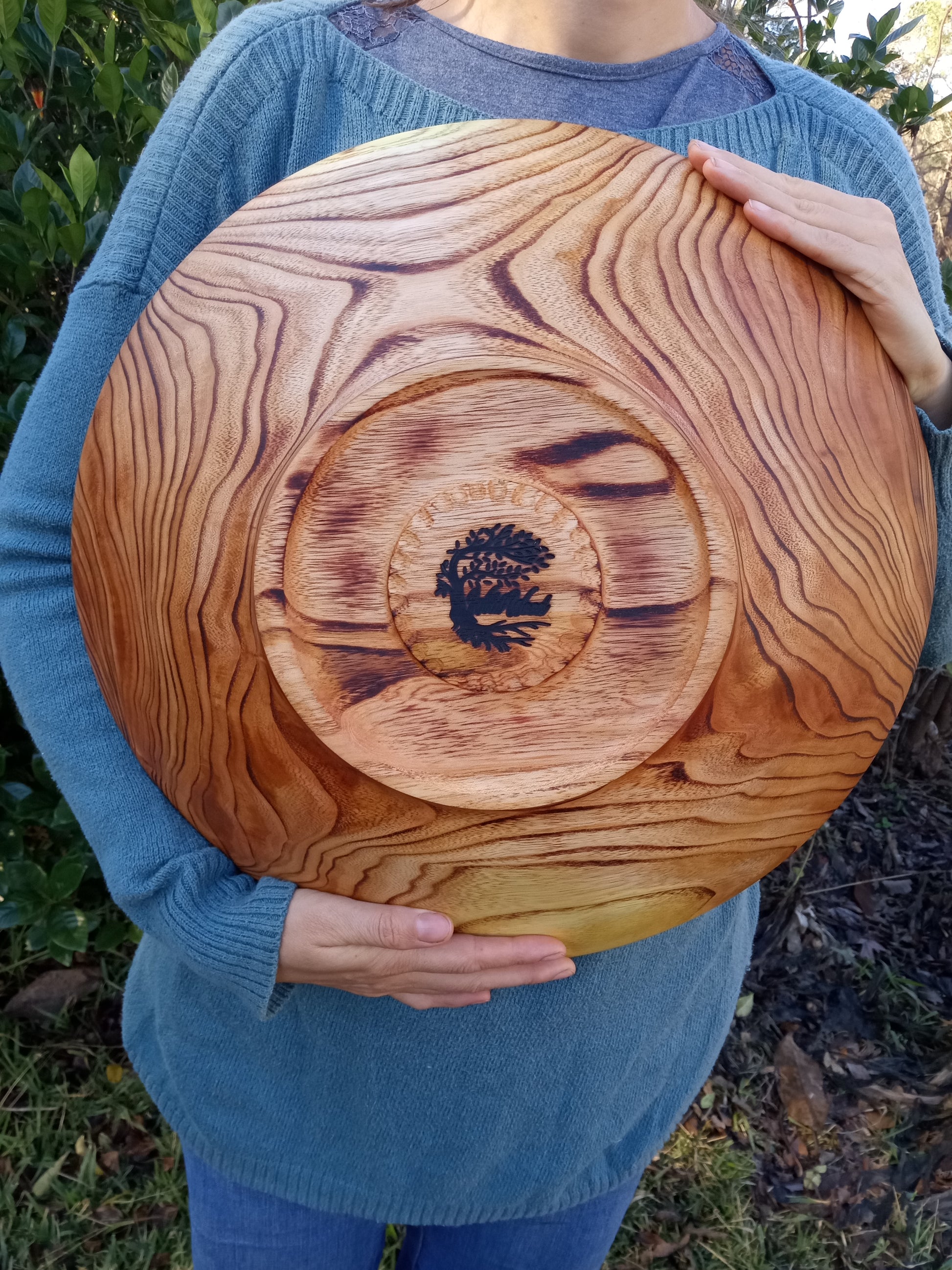 bottom view of a huge reddish toned wood bowl lightly torched on the outside to emphasize the wild grain pattern with a round footing carved into the bottom encircling a "wild wood" brand. the bowl is being held by a woman in a blue sweater