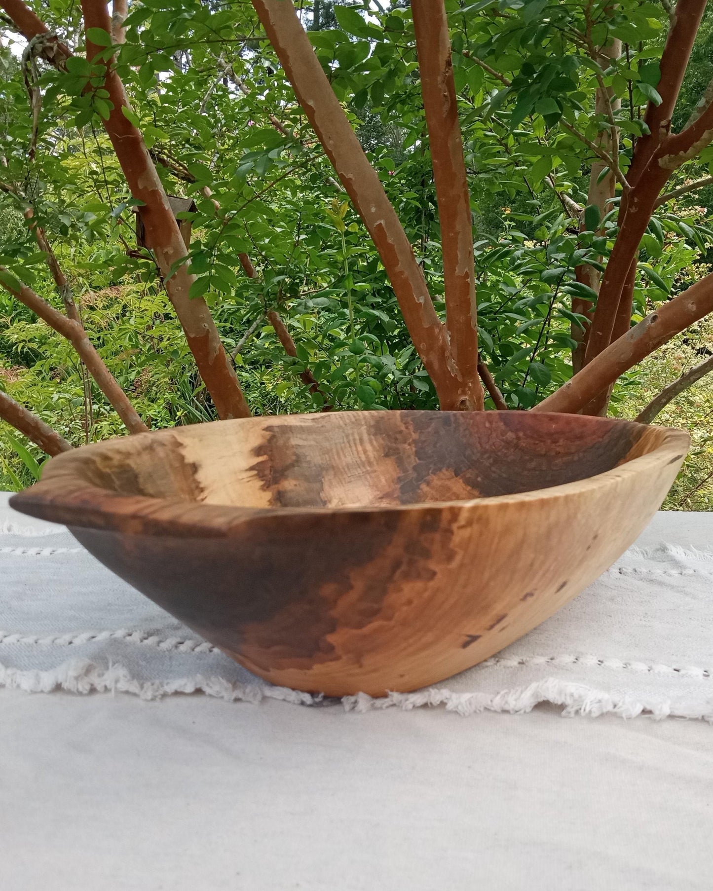 a highly figured curly maple wood dough bowl with hand carved handles and deep sidewalls set on a pale blue woven cotton table runner with tree branches in the background