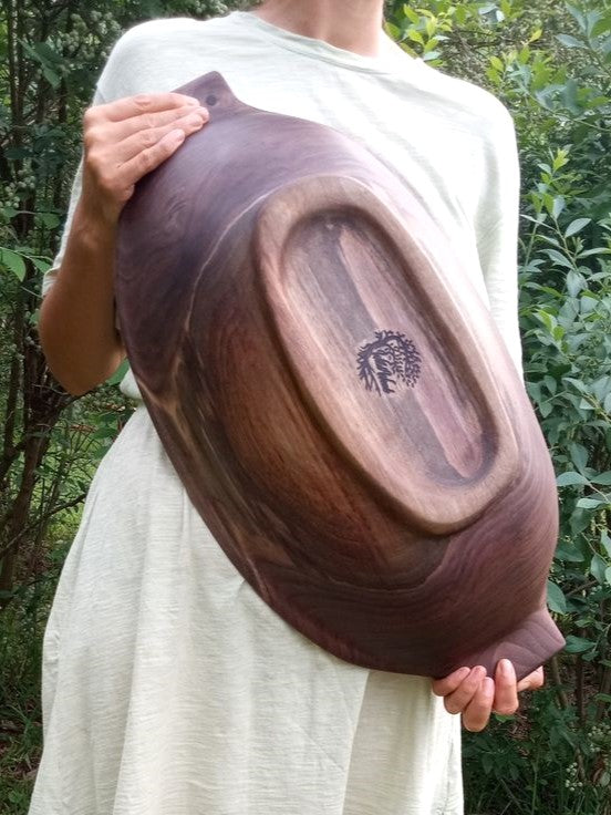 an oblong highly figured black walnut wood dough bowl with handles and a footing carved into the bottom held by a woman in a sage green t-shirt dress standing in a patch of high bush blueberries