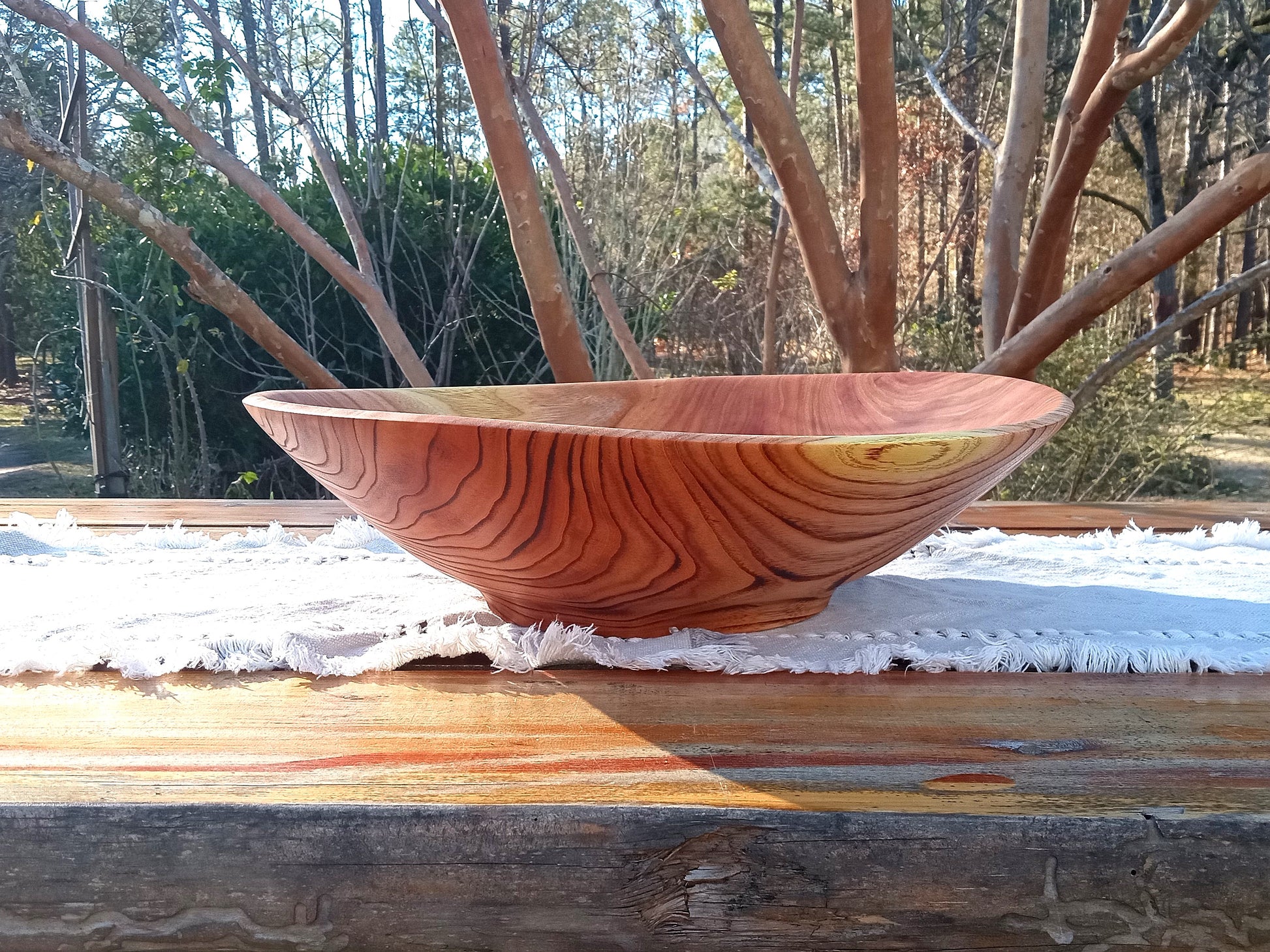 a large reddish toned wood bowl with a lightly torched outside that emphasizes the grain pattern and a  wavy natural edge set on a rustic wooden table outside