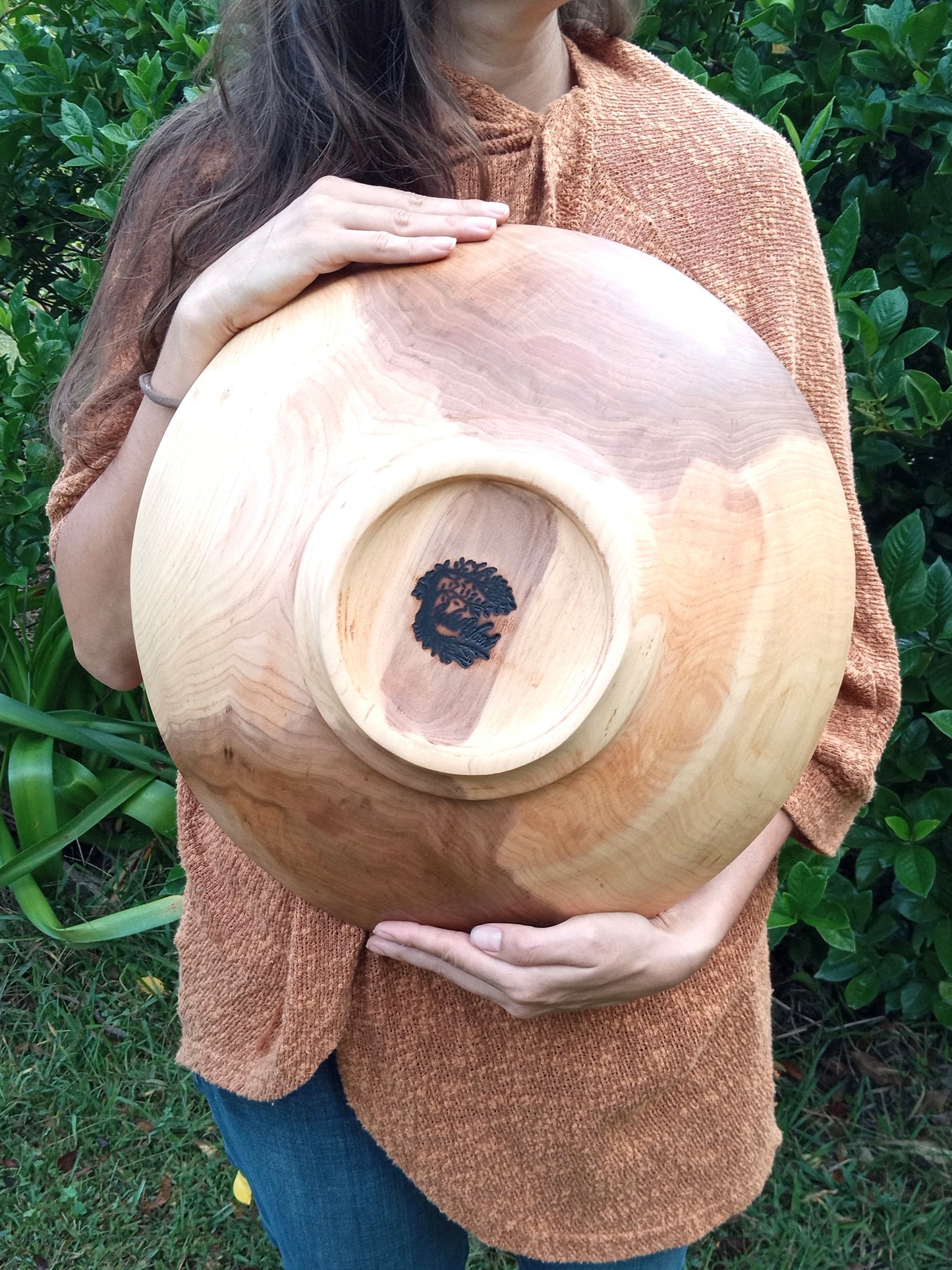 a large highly figured wood bowl with rich amber, cream and copper hues and a narrow round footing carved into the bottom encircling the Wild Wood brand held by a woman in a copper colored crochet cardigan