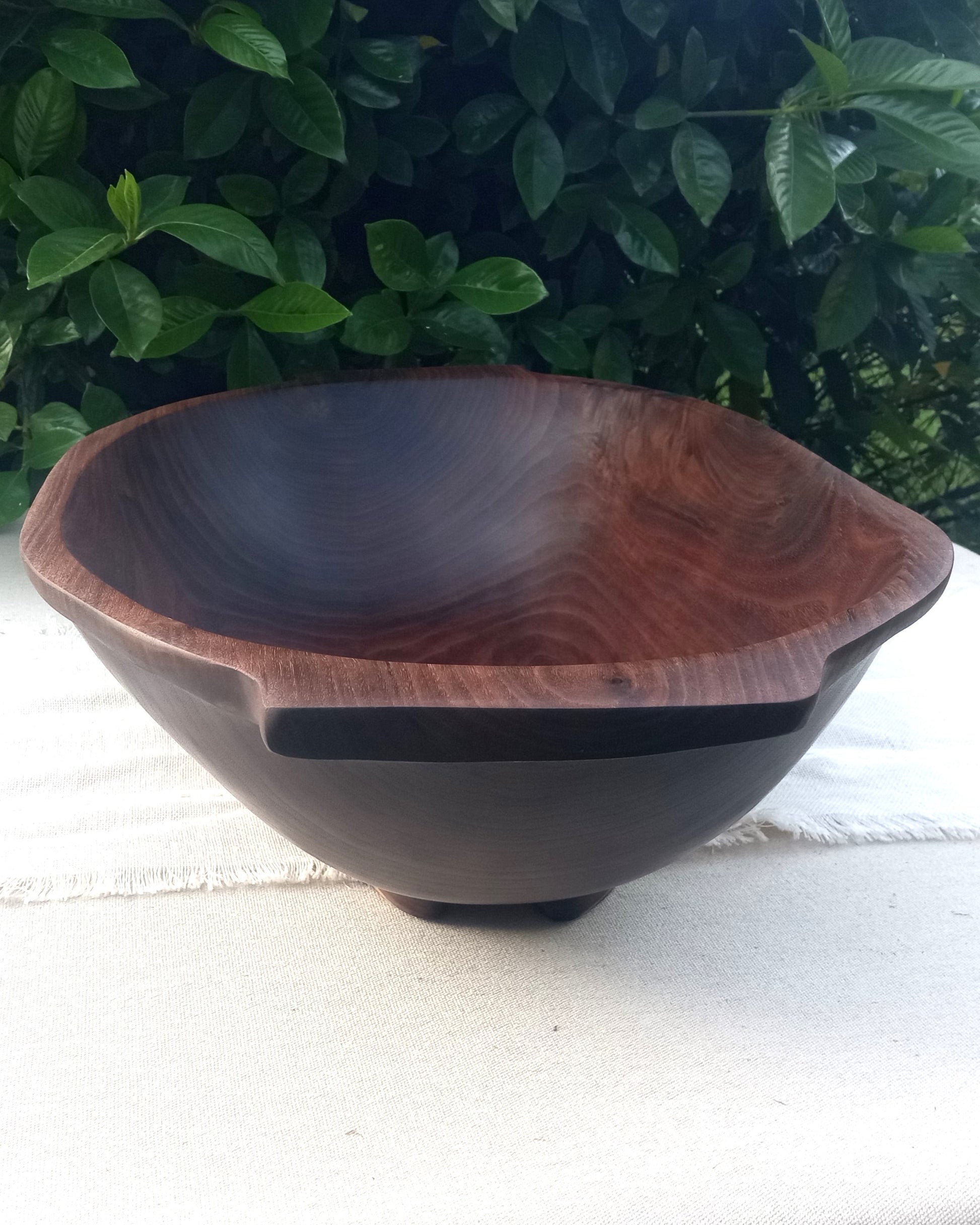 a deep oval dark walnut wood dough bowl with handles and a burl detail in the sidewall and feet carved into the bottom set on a linen tablecloth with greenery in the background